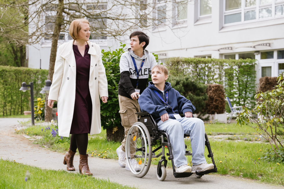 Lisa Paus mit begleitet Boys'Day-Teilnehmer im Garten der Pflegeeinrichtung, der einen Rollstuhlfahrer schiebt